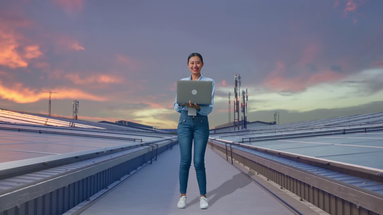 Figura intera di una donna asiatica con il suo laptop, che ispeziona pannelli solari industriali sul tetto, sta guardando la fotocamera con un sorriso.
