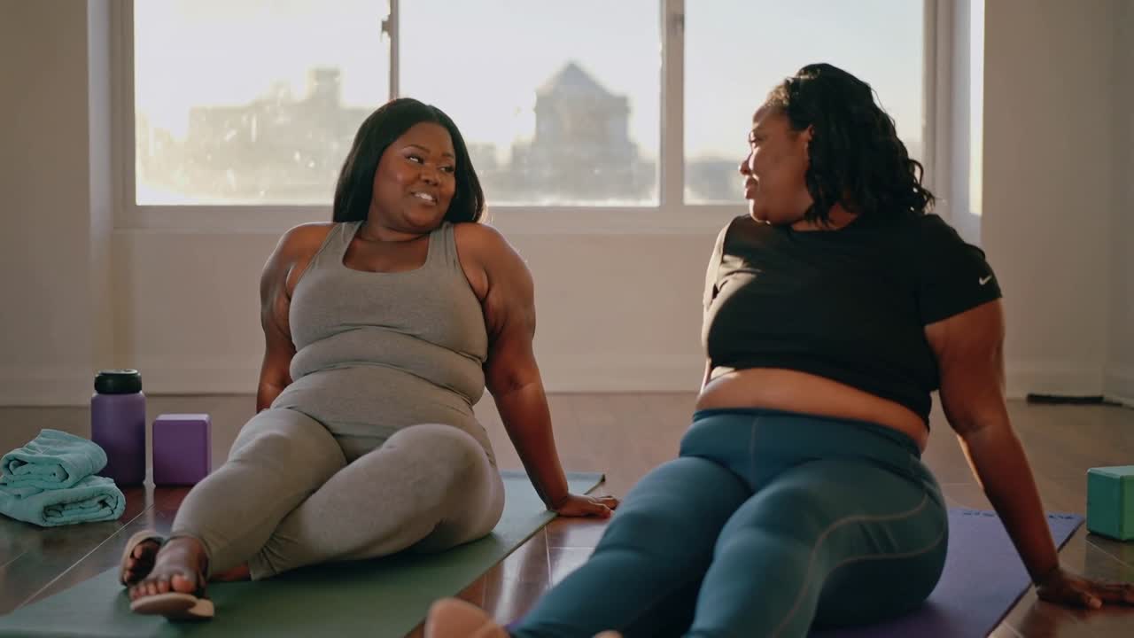 Partner sorridenti in abbigliamento sportivo grigio e nero parlano durante il defaticamento in uno studio di yoga, con tappetini.