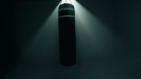 a black boxing bag gently swings left to right in a dark, smoky gym. intense backlight shines from behind, creating a glowing halo effect. scene evokes anticipation or reflection around training