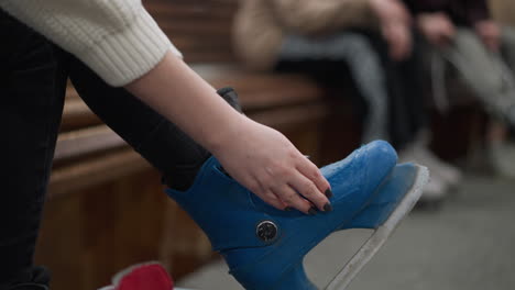 close-up of a person wearing blue ice skates and adjusting the buckles, with a focus on the hands with painted nail and black trousers, with a blur background of people