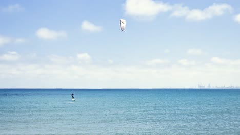 Allgemeine-Aufnahme-Einer-Person-Beim-Kitesurfen-An-Einem-Sonnigen-Tag-Mit-Blauem-Meer-Und-Einem-Weißen-Drachen-An-Einem-Windigen-Tag