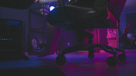 low angle leg view of woman in fishnet pantyhose walking toward gaming chair and adjusting it closer to desk in neon lit studio, with glowing equipment and mirror reflecting seating area
