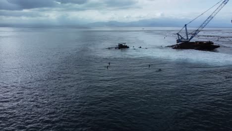 Surfers-Ride-the-Wild-Waves-Next-to-a-Crane-Shipwreck-in-Bali,-Indonesia-from-an-Aerial-Drone-Shot