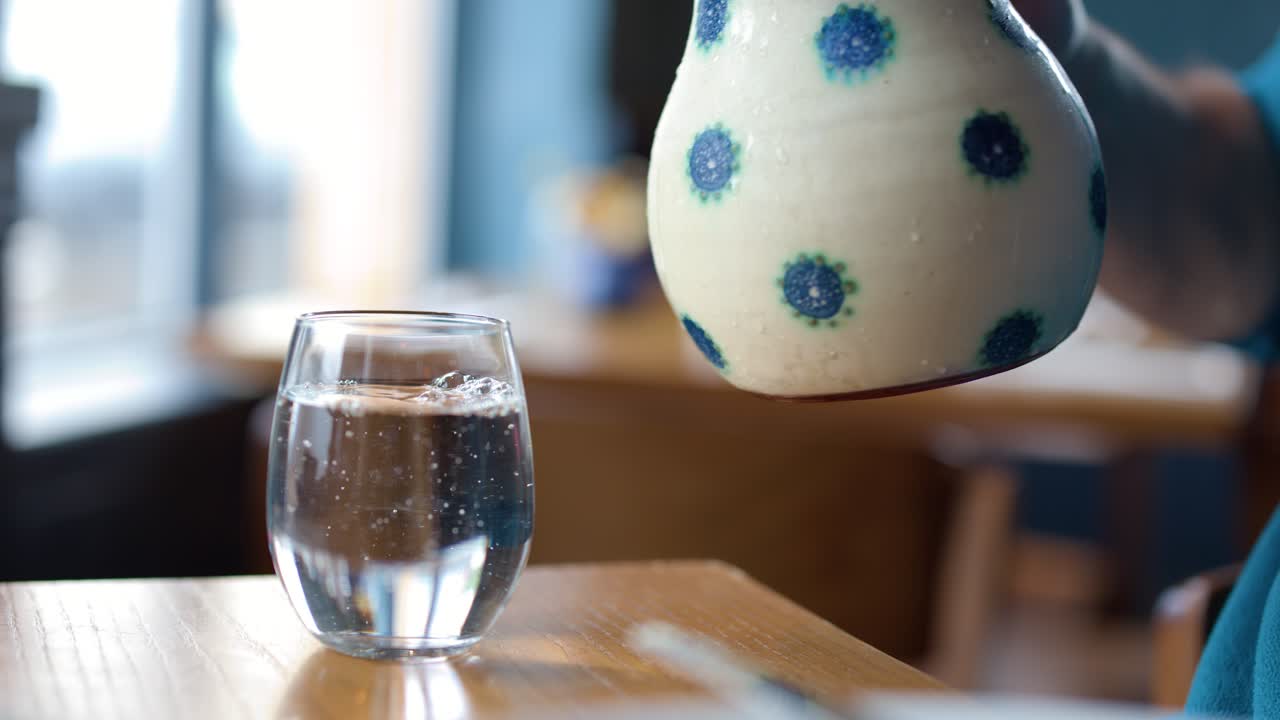 Hand pours water from patterned ceramic jug into clear glass in softly lit restaurant setting
