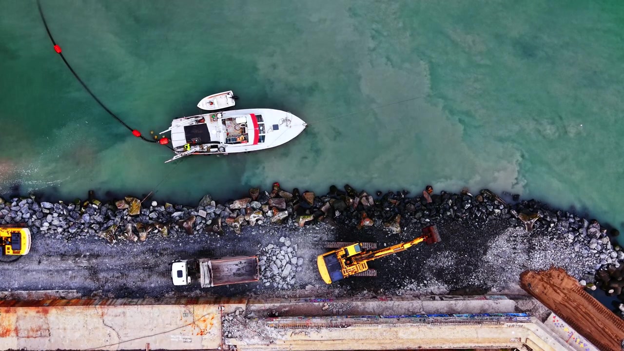 Macchinari da costruzione e imbarcazione al lavoro lungo la diga foranea del porto. Barca bianca ormeggiata accanto a un molo roccioso con due escavatori gialli e un camion sull'argine.