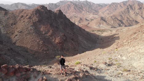 Toma-épica-Aérea-De-Un-Dron-De-Un-Joven-Parado-En-La-Cima-De-Una-Montaña-Rocosa-En-Hatta,-Emiratos-árabes-Unidos