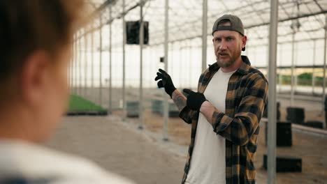 over the shoulder a confident guy in a cap with a beard in a plaid shirt communicates with a girl with red hair while designing a greenhouse and working on a farm