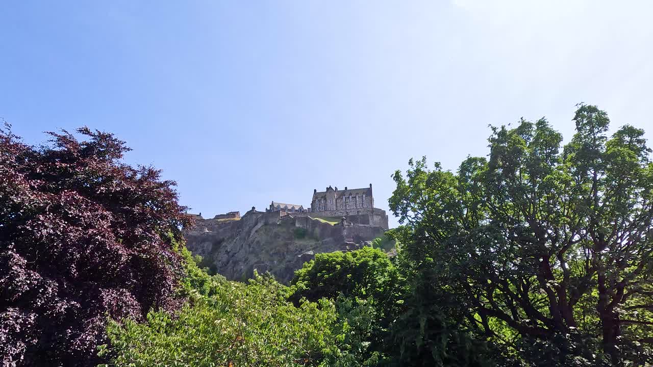 Wide shot of hilltop castle framed by green trees, under bright daylight, slight camera pan