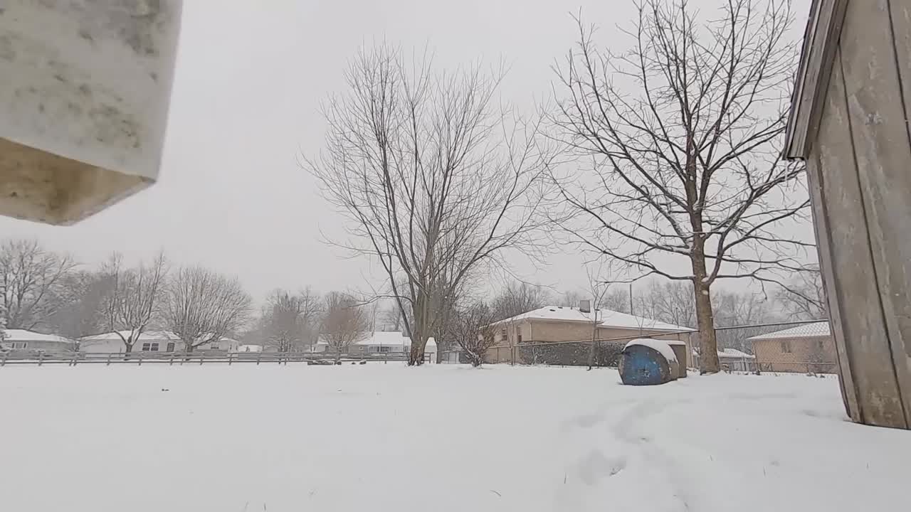 Walkthrough low to ground snow-covered gate revealing homes and a shed.