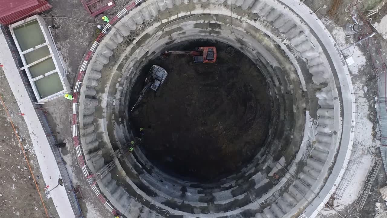 Workers climb up a lattice ladder while excavators excavate in deep tunnel shaft construction.