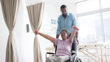 African-american-woman-exercising-with-weights-and-male-doctor-advising-in-hospital,-slow-motion