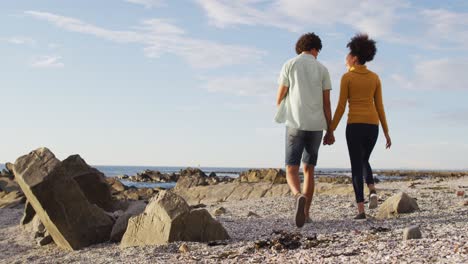 Rückansicht-Eines-Afroamerikanischen-Paares,-Das-Händchen-Hält-Und-Auf-Den-Felsen-Am-Meer-Spaziert