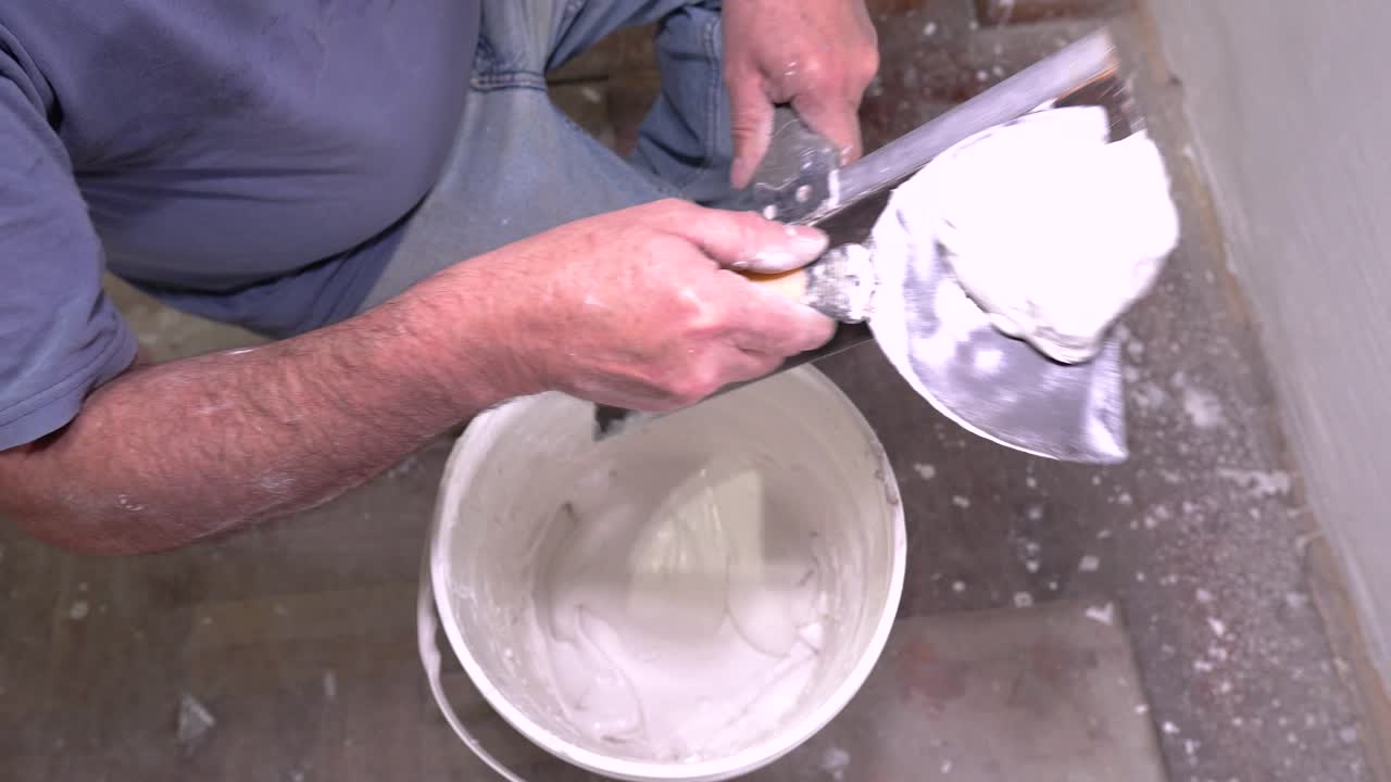 A professional worker uses a taping knife to prepare drywall mud or spackle, scooping the material from a bucket for wall finishing