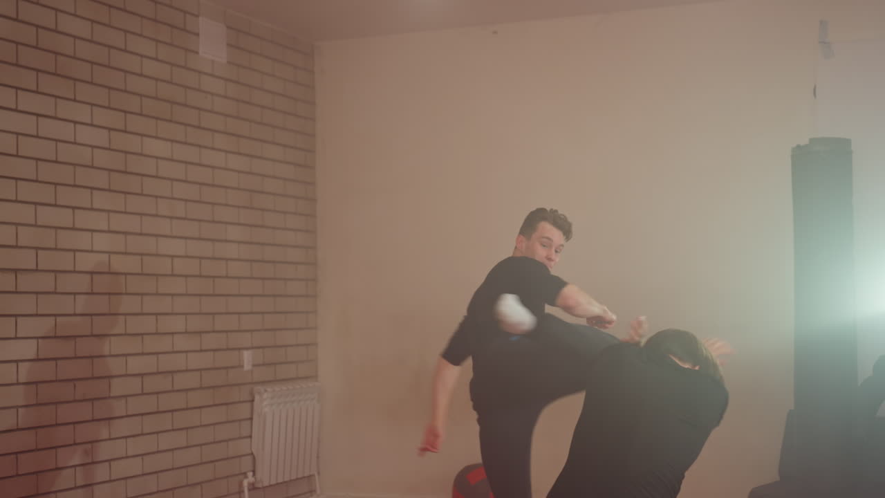 Karate practitioner performing flying strike during sparring training in gym, opponent raising arms in defense, intense martial arts action with shield impact on red mats, dynamic combat motion and energy