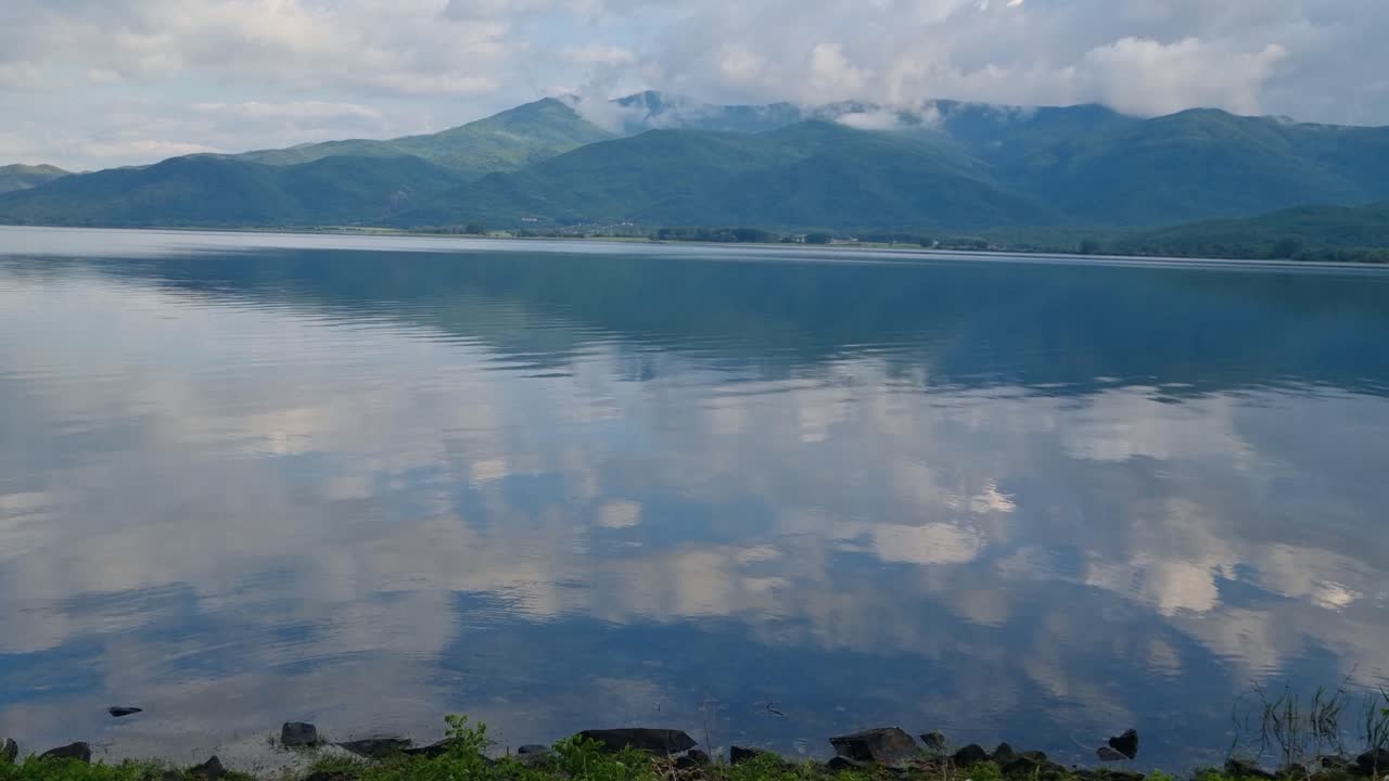 View of Kerkini Reservoir Lake in northern Greece on a cold spring day, with surrounding mountains and cloudy sky mirrored in the calm, clear water