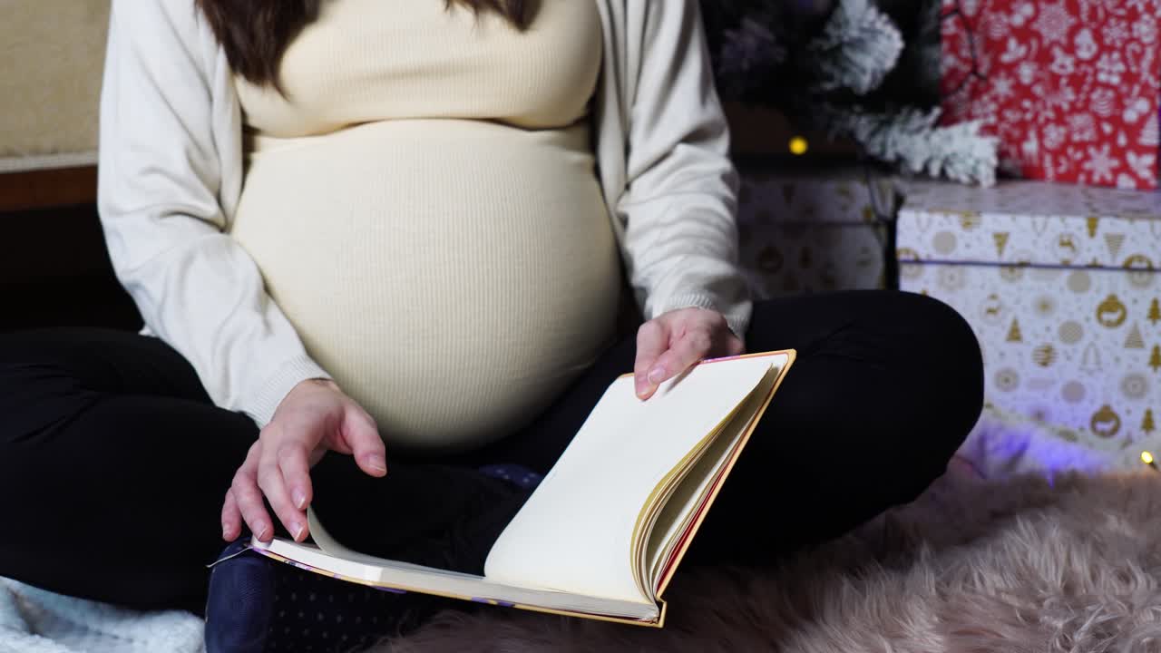 Pregnant woman sit in yoga pose and flip through notebook pages near gift boxes