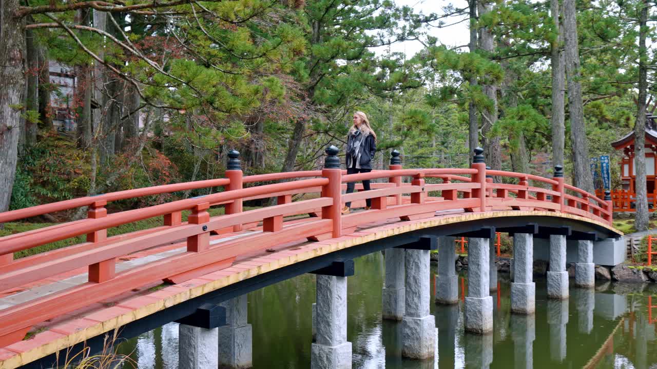 A cinematic scene capturing a blonde girl walking gracefully across a traditional bridge within the sacred grounds of Danjo Garan in Mount Koya, Koyasan, Japan.