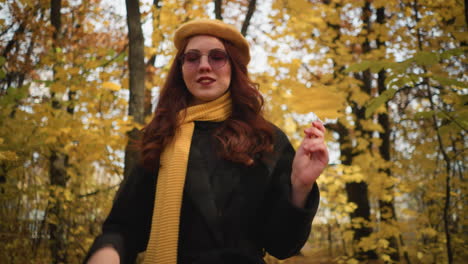 stylish lady in yellow beret and muffler playfully holds autumn leaf near face while strolling through a scenic park, surrounded by golden foliage, enjoying peaceful nature in crisp fall air