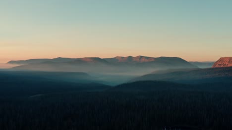 Hermosa-Toma-Aérea-De-Camiones-A-La-Izquierda-Del-Impresionante-Bosque-Nacional-Salvaje-Uinta-Wasatch-Cache-En-Utah-Con-Grandes-Pinos-Debajo-E-Impresionantes-Montañas-Cubiertas-De-Niebla-Durante-Un-Amanecer-De-Verano