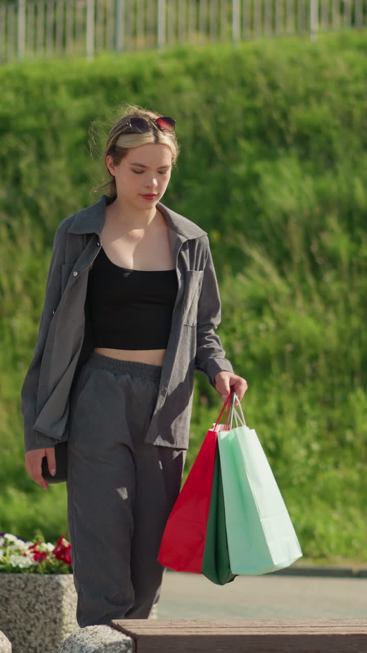 한 여인이 다채로운 쇼핑백을 들고 벤치에 다가와 앉아서 왼손을 벤치 위에 놓고 바람이 그녀의 머리카락을 흔들며 그녀 자신을 조정합니다. 배경에는 초록색과 꽃이 있습니다.