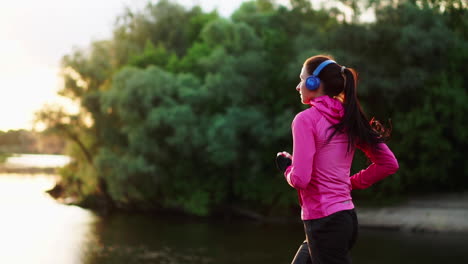 brunette with long hair in headphones runs along the river in the park in the morning at sunrise in the summer in a pink jacket and black pants