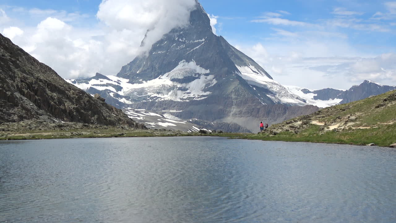 Ampla tomada da montanha Matterhorn e de um lago alpino calmo com caminhantes na margem perto de Zermatt, Valais, Suíça, sob céu azul e nuvens.