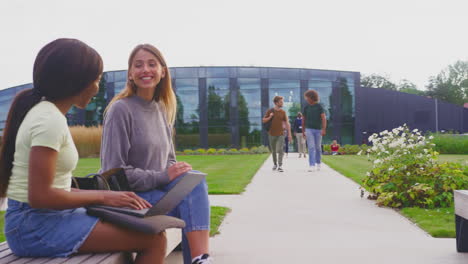 female university or college students sitting outdoors on campus talking and working on laptop