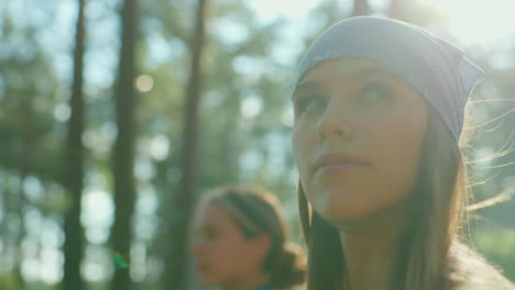 close-up of sisters walking through lush green forest, one wearing blue scarf, illuminated by sunlight reflecting off her hair as she gazes around, soft blur effect on companion