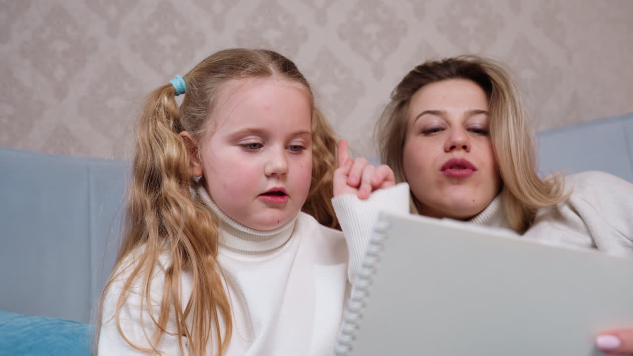 Young girl with long blonde hair and woman in cozy sweaters sitting on couch looking at sketchbook together, sharing quiet creative bonding time with drawing activity in warm relaxed home environment