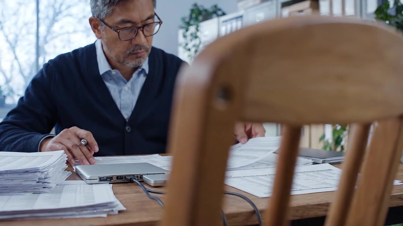 Uomo concentrato che lavora su documenti con laptop e documenti in un ambiente d'ufficio luminoso, circondato da schedari e con luce naturale che entra.