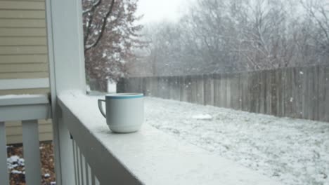 Eine-Tasse-Heißen-Kaffee-Steht-Auf-Einem-Balkongeländer,-Während-Im-Winter-Im-Hinterhof-Eines-Bauernhauses-In-Zeitlupe-Schnee-Fällt