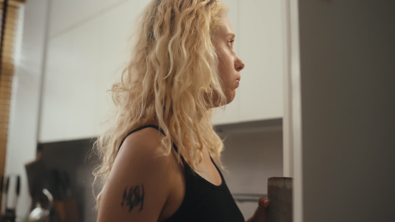 Rear view of a blonde girl with curly hair drinking coffee and looking into the refrigerator with a phone in her hands in the morning