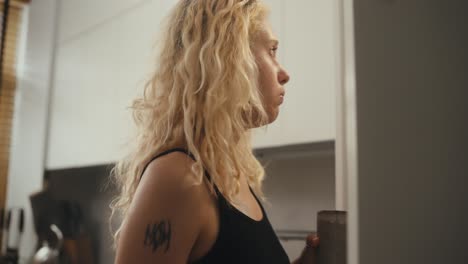 Rear-view-of-a-blonde-girl-with-curly-hair-drinking-coffee-and-looking-into-the-refrigerator-with-a-phone-in-her-hands-in-the-morning