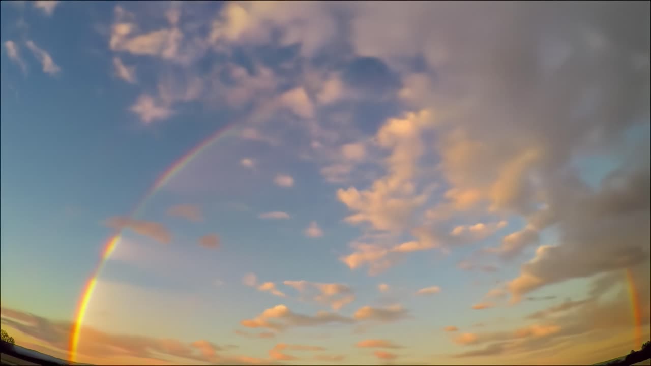 A brilliant rainbow stretches across a colorful sky filled with soft clouds as the sun sets. The scene captures the beauty of nature in a serene location during twilight.