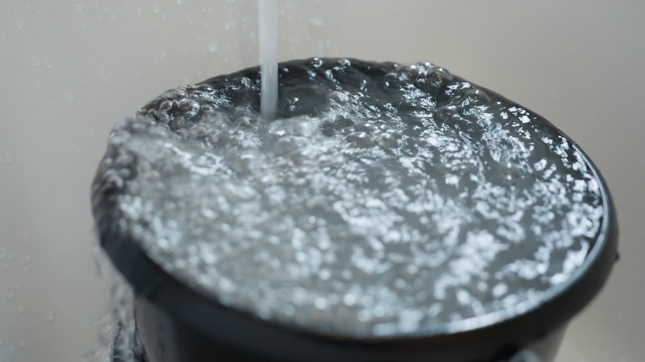 Close up of water running into black bucket filled to brim, liquid bubbling and pouring out over edge, illustrating overflow, home cleaning, water use, sanitation, daily utility task and fluid movement