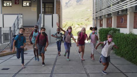 Diverse-group-of-schoolchildren-wearing-backpacks-and-face-masks-running-at-school-yard