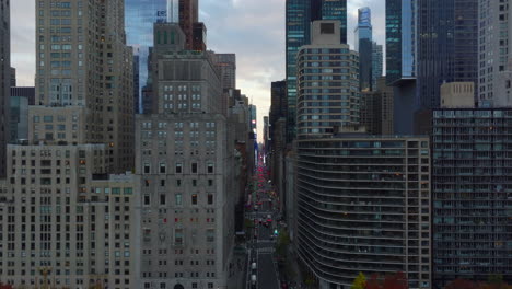 Forwards-fly-into-space-between-tall-buildings-encircling-Central-park.-Fly-above-wide-avenue,-red-brake-lights-of-cars.-Manhattan,-New-York-City,-USA