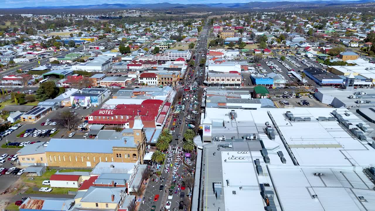 Drone footage glides above a bustling main street lined with crowds, colorful buildings, and festival activity on a clear, sunny day