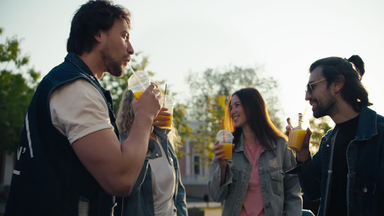 un grupo de jóvenes en chaquetas de vaqueros de moda sonan sus vasos de plástico con un cóctel amarillo en la calle en un día soleado