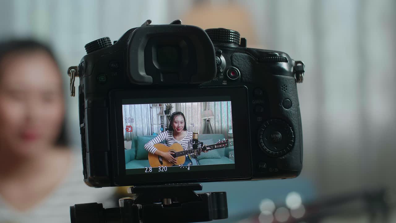 close up de um monitor de câmera gravando mulher asiática agitando a mão e falando antes de tocar guitarra em casa