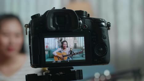 cerca de un monitor de cámara grabando a una mujer asiática agitando la mano y hablando antes de tocar la guitarra en casa