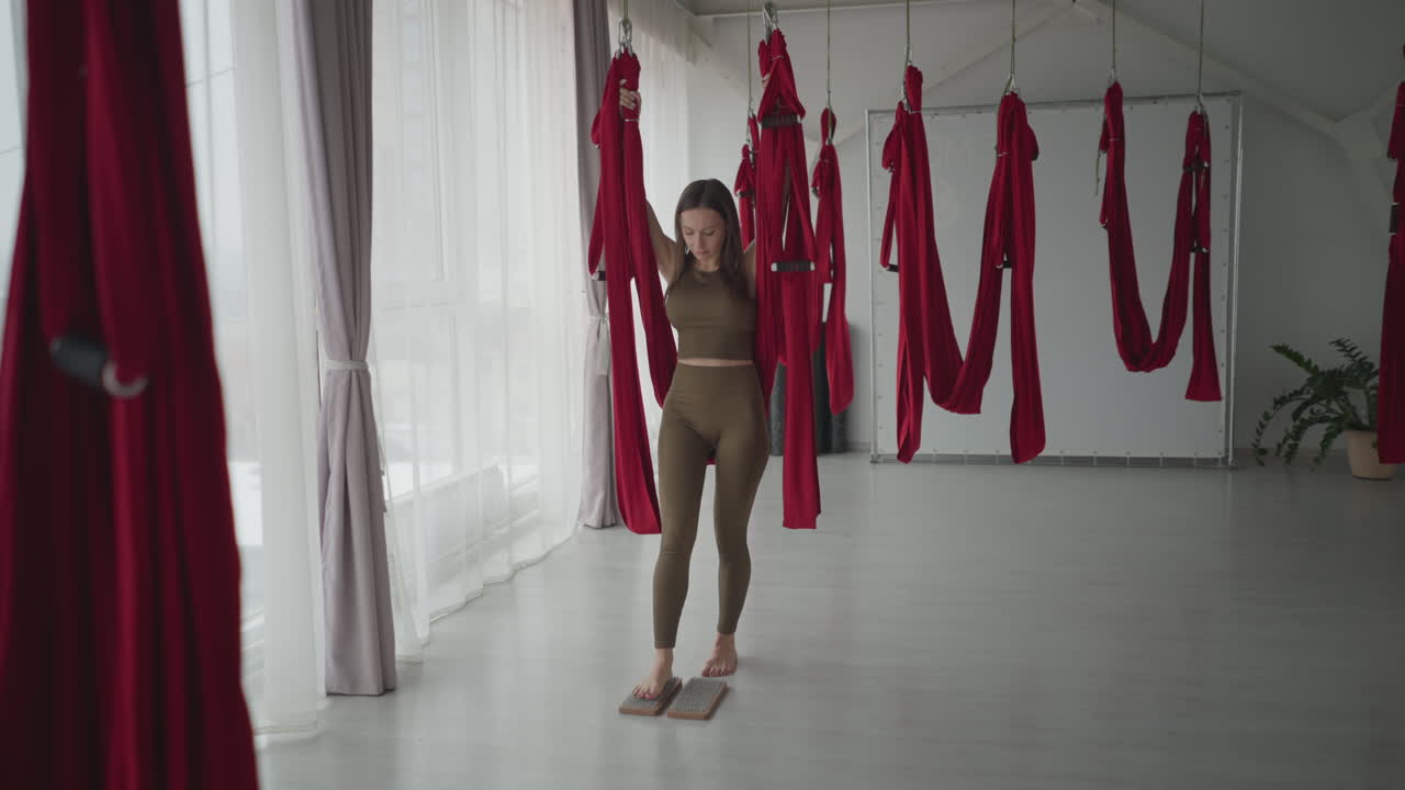 Fitness coach in olive activewear balances barefoot on wooden nail blocks while gripping red aerial silk hammock inside bright studio with floor to ceiling windows and suspended silks