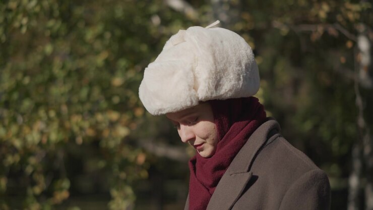 Primo piano di una donna che indossa abiti invernali tradizionali, inclusi un cappello di pelliccia bianco e una sciarpa bordeaux, in piedi all'aperto con luce naturale. Il soggetto rimane immobile per tutta la scena con una luce costante.
