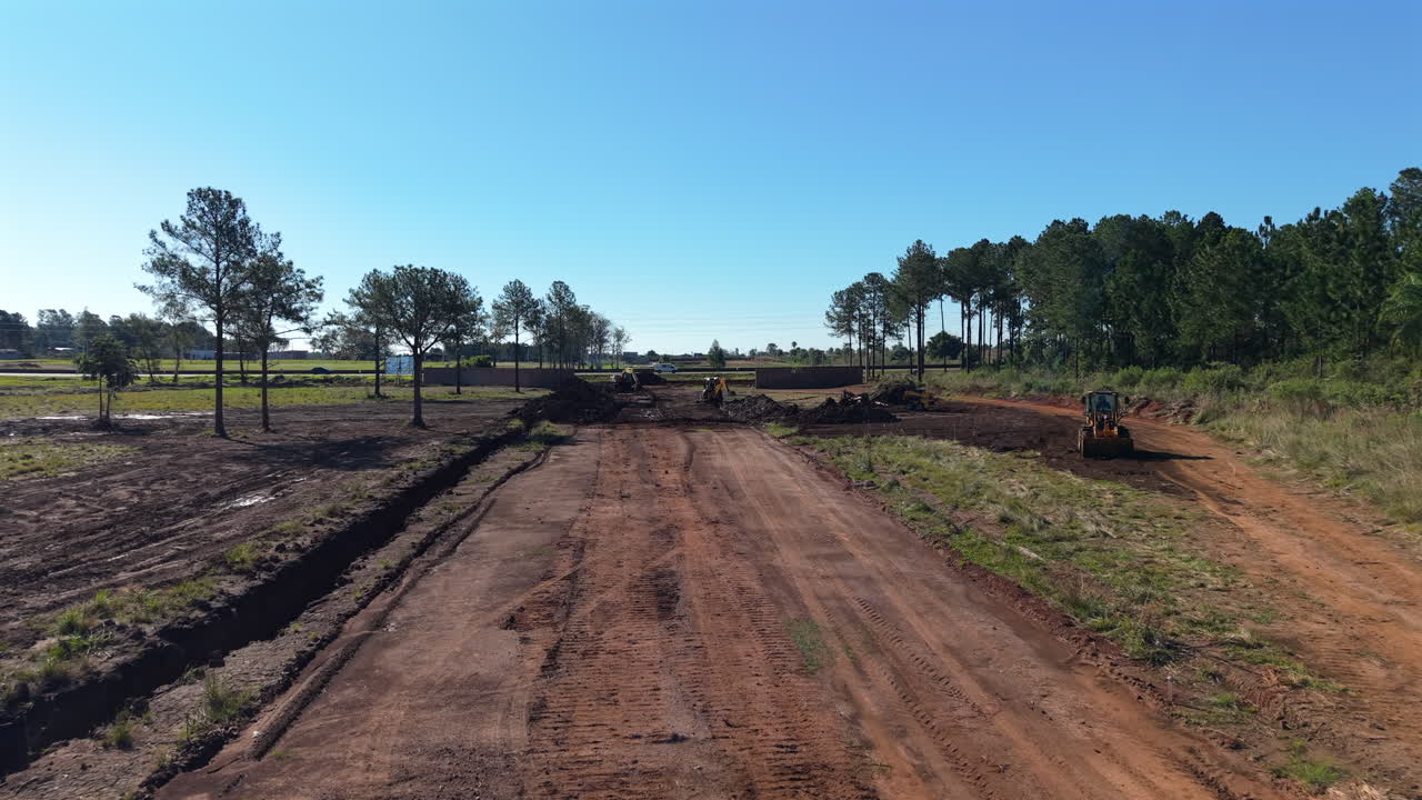 Construction site roadwork clearing, wide sunny view with trees and machinery