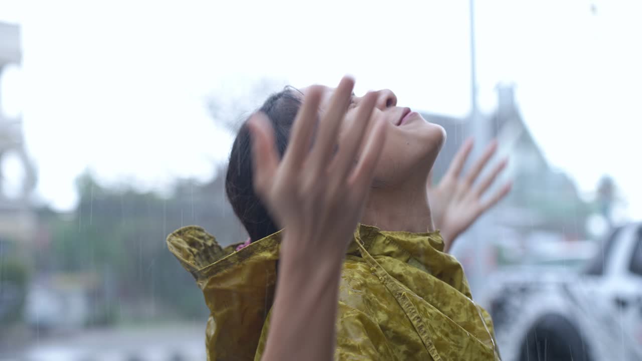 아시안 소녀는 방울 코트를 벗고 아오르는 비를 즐기기 위해 도로 에 서서 하늘을 쳐다보고 얼굴에 비가 떨어지는 것을 즐기고 미소 짓고, 신선함을 느끼고, 계절, 기후 변화를 느니다.