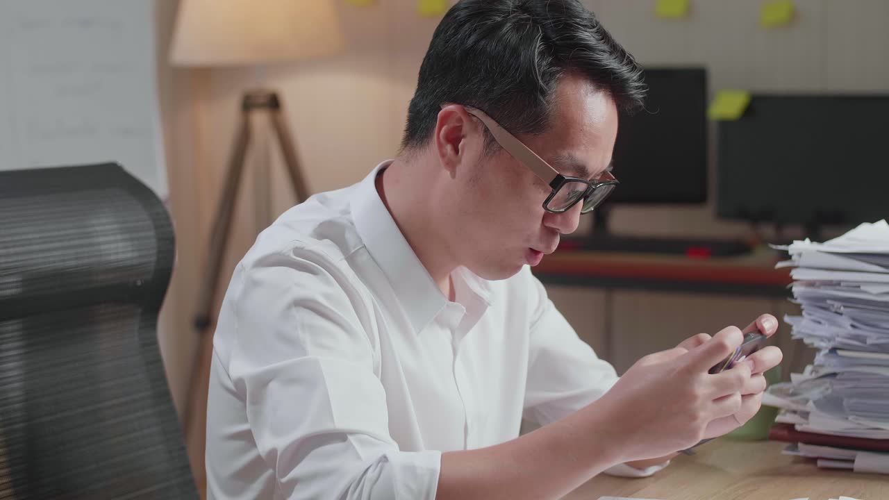 행복한 아시아 남자가 사무실에서 문서와 함께 일한 후 스마트폰에서 우승 게임을 축하하는 클로즈업