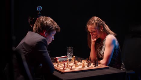 man and woman compete in serious chess tournament match, man captures white piece with trophy in background- low light clip