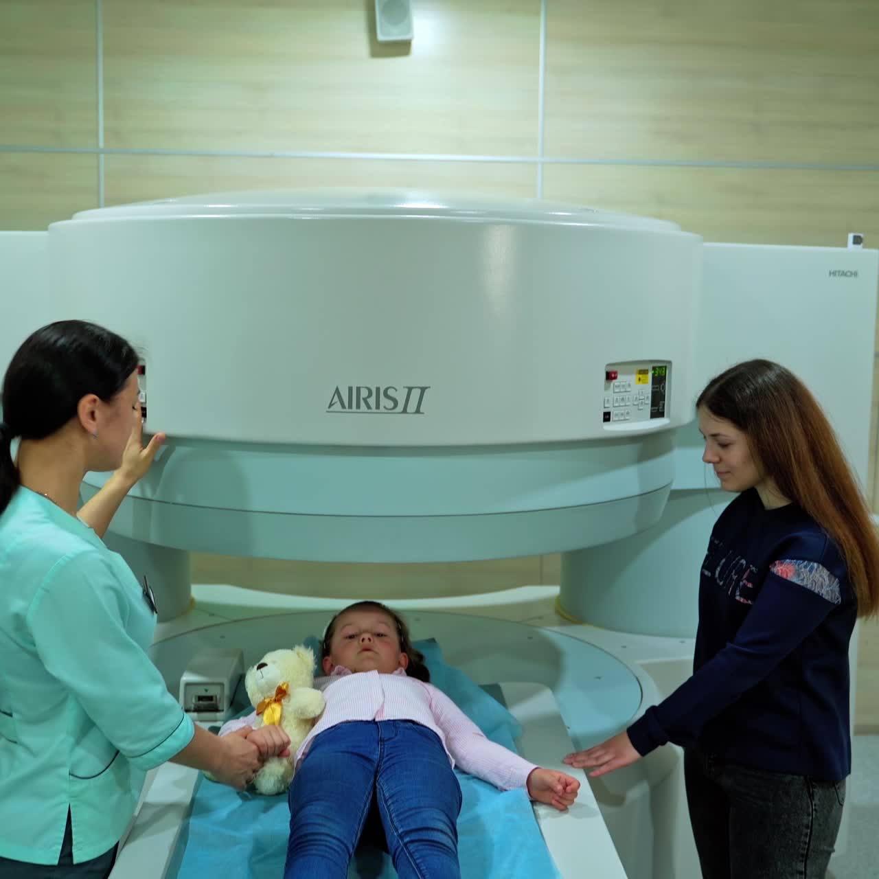 Una bambina è sdraiata nel macchinario per la risonanza magnetica in un centro medico. Un medico e la madre sono in piedi su entrambi i lati della bambina.