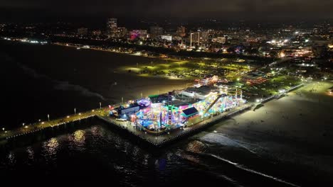 rising above santa monica pier at night, aerial of amusement park of rides lit up in the dark