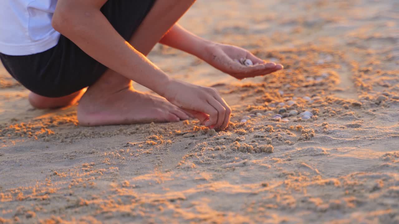 Boy is playing on sand beach at sunset. Boy making something with small pebbles on sand in the evening. Happy childhood in summer time.